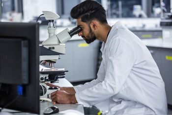 A male biomedical scientist working in a lab, looking through a microscope.